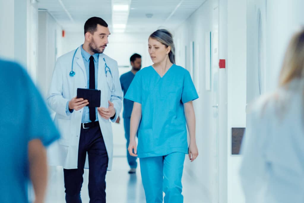 Surgeon and Female Doctor Walk Through Hospital Hallway, They Consult Digital Tablet Computer while Talking about Patient's Health. Modern Bright Hospital with Professional
